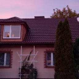 Dom jednorodzinny z brązowym dachem z dachówki bitumicznej, widoczny lukarna i komin na tle różowego nieba o zmierzchu.