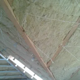 Poddasze z ociepleniem z wełny mineralnej, widoczne drewniane krokwie i metalowe profile, całość zabezpieczona sznurkiem.