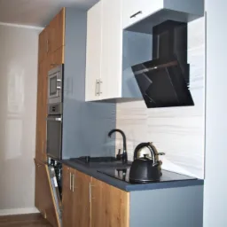 Nowoczesna kuchnia z drewnianymi szafkami dolnymi, białymi szafkami g&oacute;rnymi i szarym blatem. Widoczny czarny okap, zlew i czajnik elektryczny. Otwarta zmywarka. Podłoga wyłożona panelami...