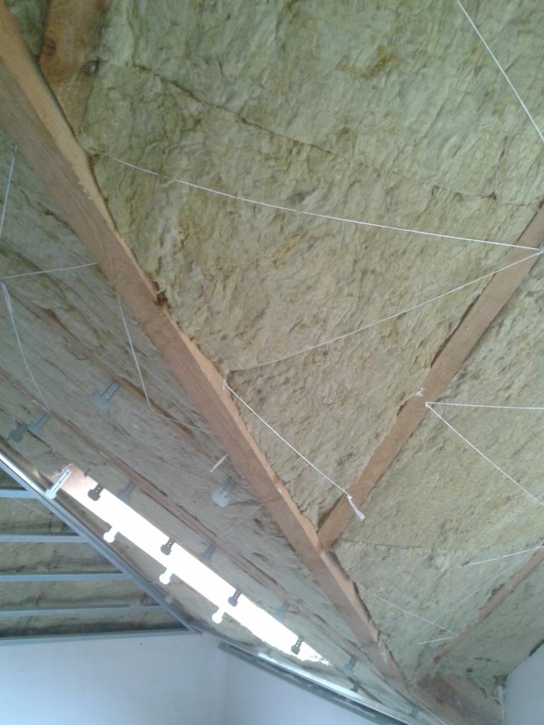 Poddasze z ociepleniem z wełny mineralnej, widoczne drewniane krokwie i metalowe profile, całość zabezpieczona sznurkiem.