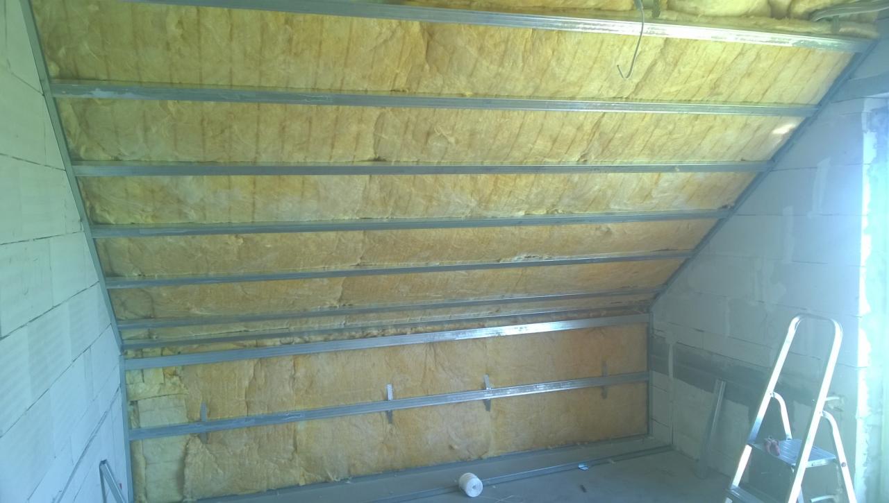 Ocieplenie poddasza wełną mineralną, widoczne metalowe profile konstrukcyjne i fragment ściany z bloczków, drabina aluminiowa w rogu.