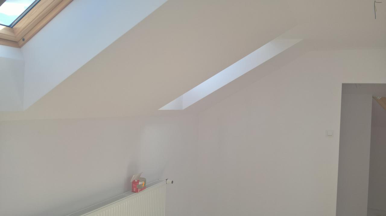 Poddasze po remoncie, z oknem dachowym i gładkimi, pomalowanymi na jasny kolor ścianami. Widoczny grzejnik i pudełko chusteczek na nim.