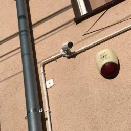 Kamera monitoringu i sygnalizator alarmowy zamontowane na elewacji budynku, widoczny rynna spustowa. Instalacja wykonana z rur PCV.