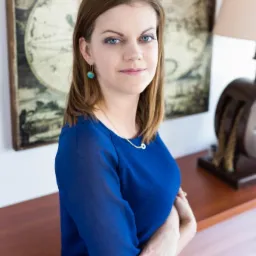 Portret kobiety w niebieskiej bluzce na tle mapy świata i lampy na drewnianym biurku.