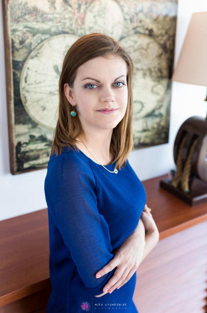 Portret kobiety w niebieskiej bluzce na tle mapy świata i lampy na drewnianym biurku.