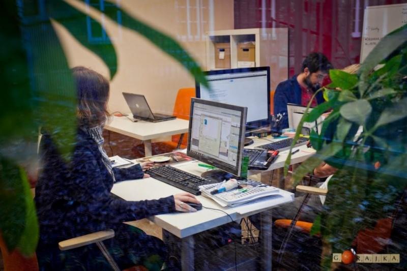 Wnętrze biura z dwoma pracownikami przy biurkach, widoczne przez przeszklenie z roślinami, jeden pracownik używa komputera stacjonarnego, drugi laptopa, na biurkach leżą dokumenty i akcesoria biurowe.