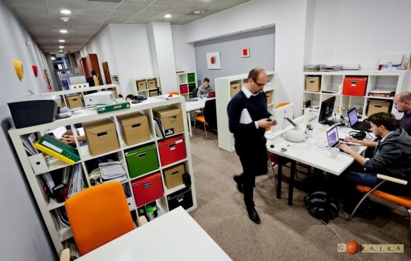 Przestronne biuro z pracownikami przy biurkach, regały z kolorowymi pojemnikami i mężczyzna w czarnym swetrze używający smartfona.