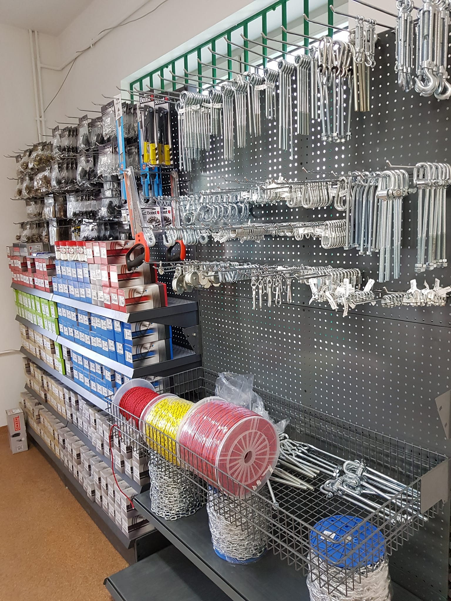 Ekspozycja różnorodnych materiałów i akcesoriów: liny w kolorach czerwonym i żółtym, łańcuchy, haki, śruby, nakrętki i inne elementy metalowe, rozmieszczone na półkach i perforowanej ścianie...