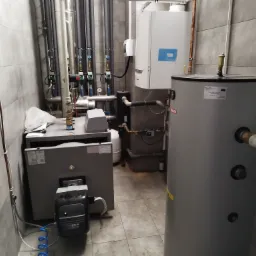 Szara kotłownia z piecem, zasobnikiem ciepłej wody i rurami. Zastosowano szare płytki na ścianach i podłodze. Widoczne zawory i manometry.