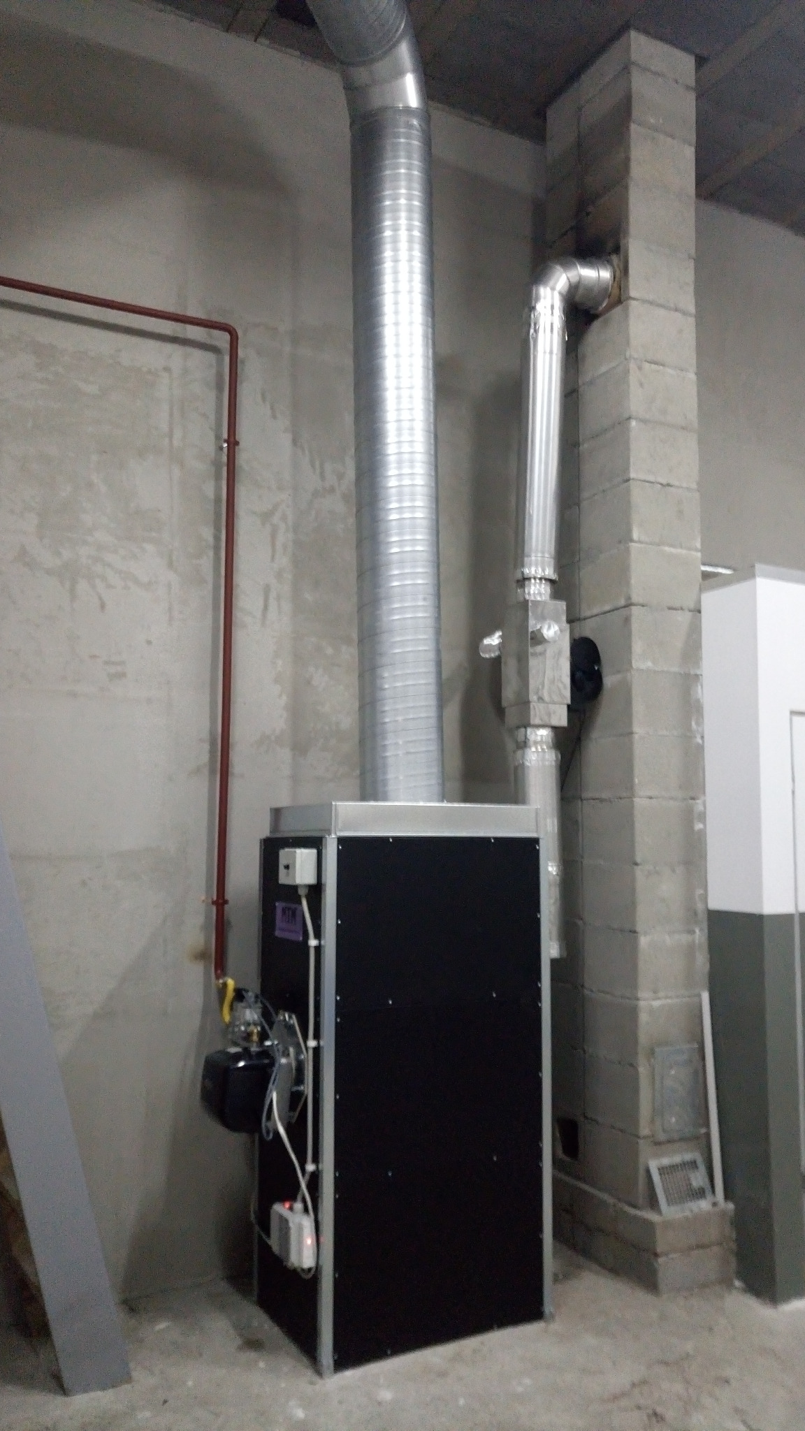 Czarny piec z metalowymi rurami wentylacyjnymi, zainstalowany w surowym pomieszczeniu z betonowymi ścianami i podłogą. Widoczne elementy instalacji i komin.