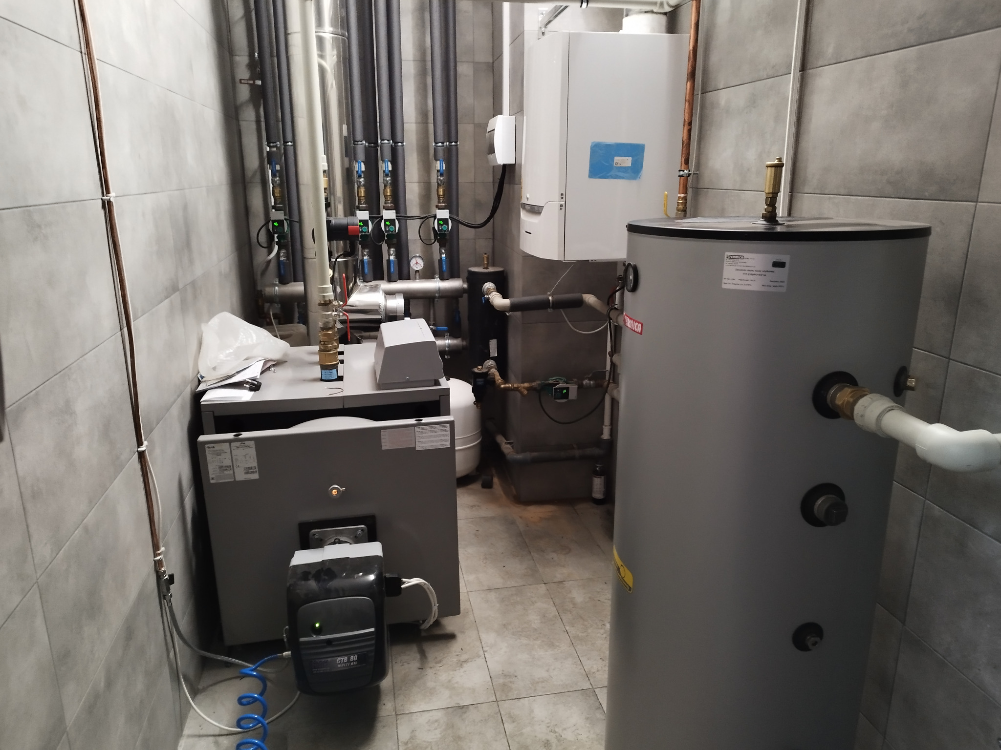 Szara kotłownia z piecem, zasobnikiem ciepłej wody i rurami. Zastosowano szare płytki na ścianach i podłodze. Widoczne zawory i manometry.