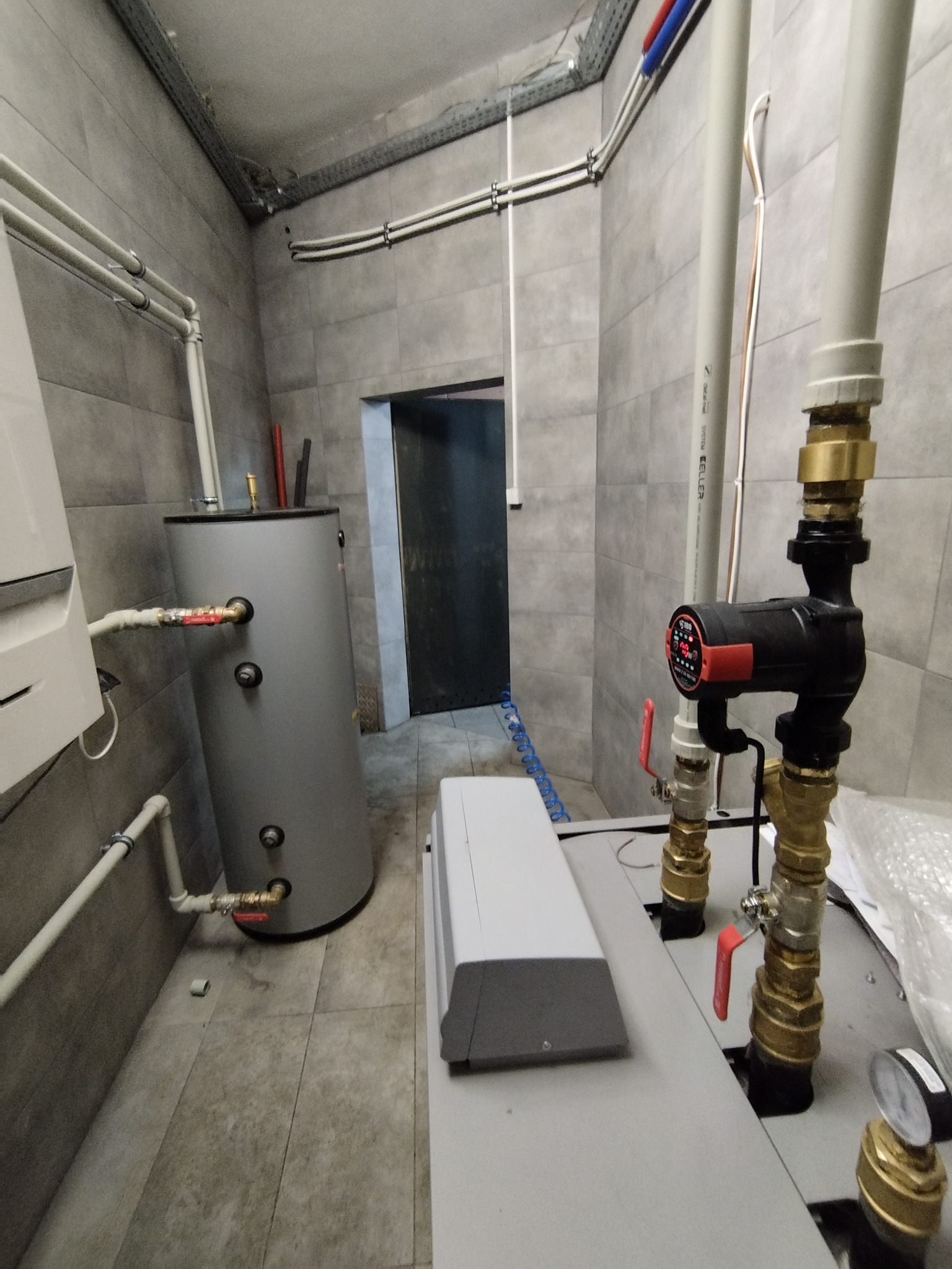 Szara kotłownia z bojlerem, pompą obiegową i instalacją w szarych płytkach. Widoczne rury, zawory i elementy automatyki. Ujęcie z dołu.