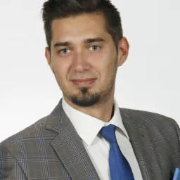 Portret młodego mężczyzny w szarej marynarce w kratę i niebieskim krawacie, z poszetką w kieszeni, na białym tle.