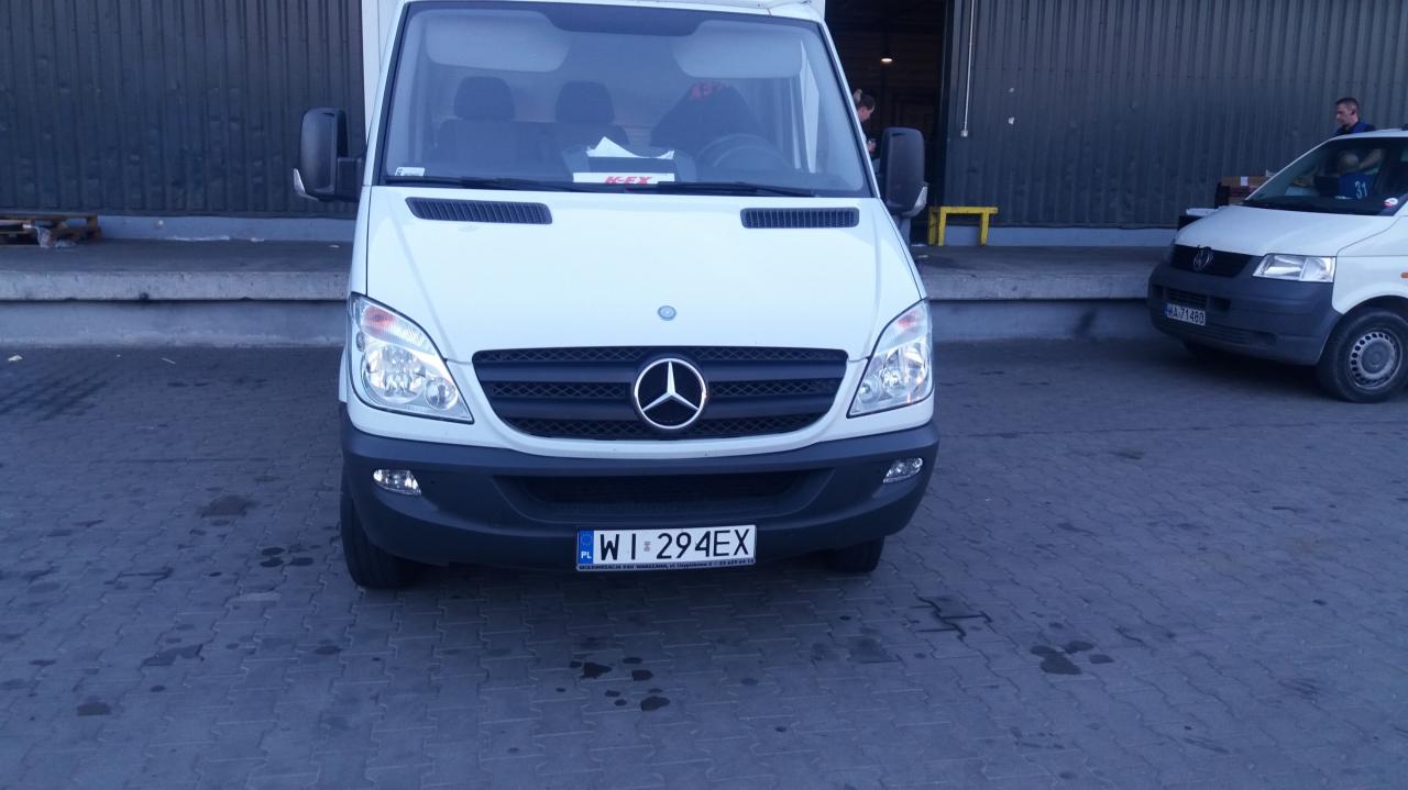 Biały bus marki Mercedes-Benz Sprinter z widoczną tablicą rejestracyjną WI 294EX, zaparkowany na zewnątrz, zarys budynku w tle.