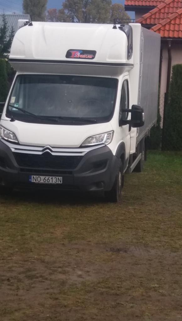 Biały samochód dostawczy z plandeką firmy z Warszawy, zaparkowany na trawiastym podwórku przed domem.