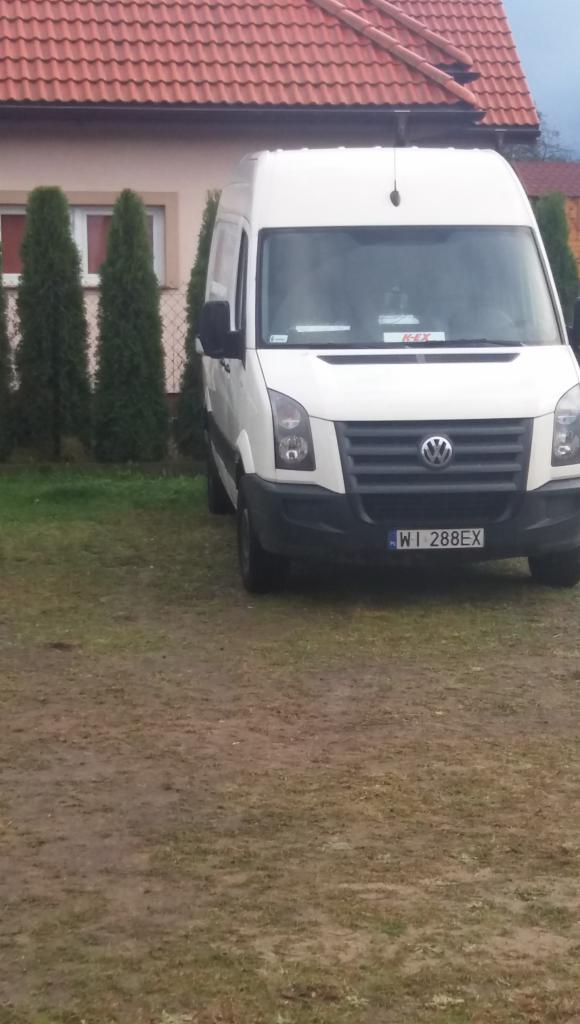 Biały van Volkswagen Crafter zaparkowany na trawniku przed domem z czerwoną dachówką, widok z przodu.