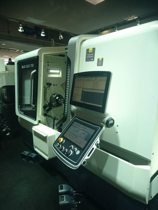 Nowoczesna, biała tokarka CNC NLX 2500|700 z otwartą komorą roboczą i dotykowym panelem sterowania z przyciskami i pokrętłami, prezentowana na hali wystawienniczej.