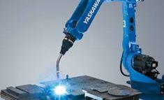 Niebieski robot spawalniczy marki Yaskawa w trakcie pracy, iskry i światło spawania widoczne na metalowej powierzchni, neutralne szare tło.