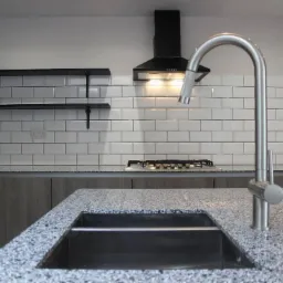 Nowoczesna kuchnia z szarymi szafkami, blatem z szarego granitu, białymi płytkami metra z czarną fugą i chromowaną baterią kuchenną skierowaną w stronę czarnego zlewu.