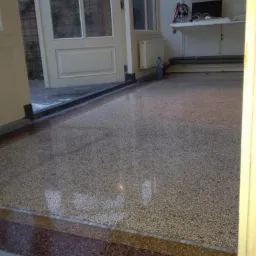 Błyszcząca, wielobarwna posadzka lastryko w pomieszczeniu z otwartymi drzwiami na zewnątrz, widoczny fragment p&oacute;łki z elektroniką i butelka wody mineralnej przy grzejniku.