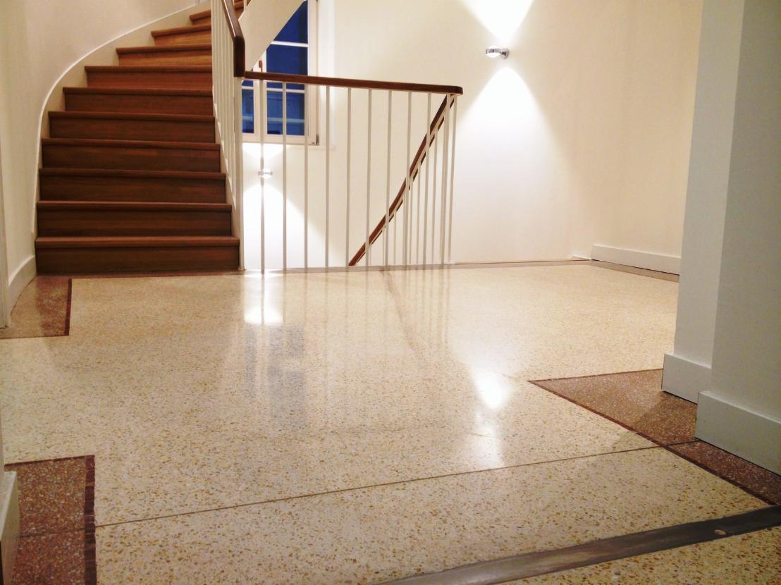 Polerowana posadzka lastryko w holu z drewnianymi schodami i białymi balustradami, widoczne detale wykończenia podłogi w kontrastowym kolorze.