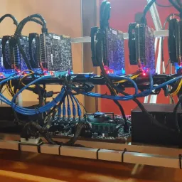 Rzędowy układ kart graficznych z chłodzeniem, zamontowanych na stelażu z aluminiowych profili, z widocznym okablowaniem i płytą główną w tle, na biurku w pomieszczeniu z czerwonymi ścianami.