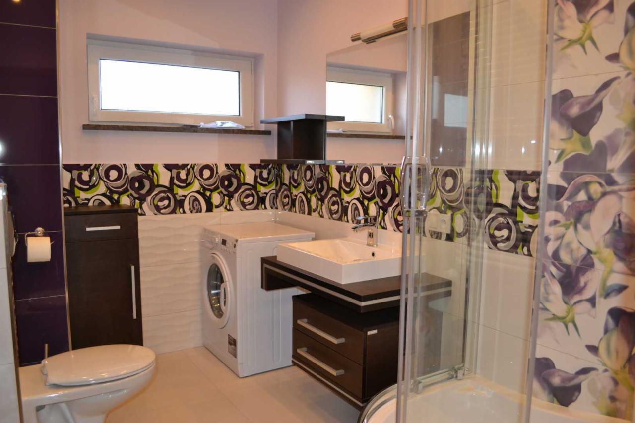 Nowoczesna łazienka z białą pralką, umywalką na szafce w kolorze ciemnego drewna, toaletą i kabiną prysznicową, płytki z motywem kwiatowym i geometrycznym.