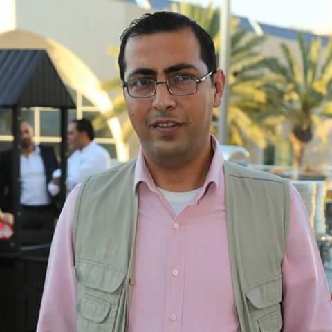 Portret mężczyzny w okularach, w różowej koszuli i beżowej kamizelce na tle palm i jasnego budynku. Uśmiechnięty, patrzy w obiektyw. W tle widoczne inne osoby.