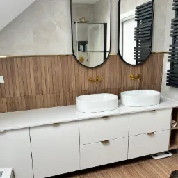 Nowoczesna łazienka na poddaszu z drewnianą boazerią, dwoma owalnymi umywalkami nablatowymi z złotą armaturą, czarnymi owalnymi lustrami i grzejnikiem drabinkowym w kolorze czarnym.