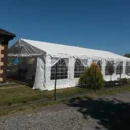 Długi, biały namiot imprezowy z oknami, ustawiony na trawniku obok domu jednorodzinnego, ogrodzony siatką.