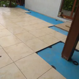 Układanie beżowych płytek na tarasie z widocznymi podkładkami dystansowymi i niebieską folią izolacyjną.