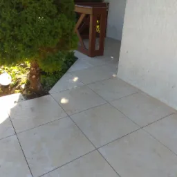 Jasne, kwadratowe płytki podłogowe na zewnątrz, z zielonym krzewem w rogu i fragmentem drewnianej konstrukcji.