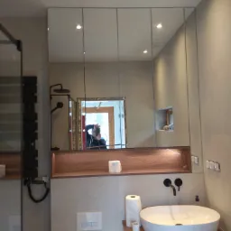 Nowoczesna łazienka z umywalką nablatową, lustrem szafkowym z trzema segmentami i drewnianą półką z oświetleniem LED, widoczne odbicie fotografa w lustrze.