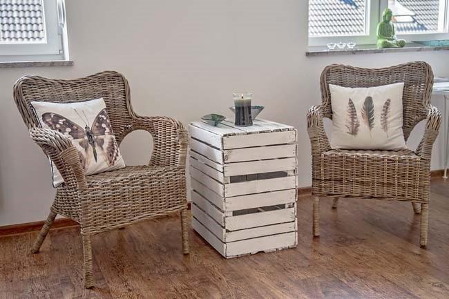 Jasne, minimalistyczne wnętrze z dwoma wiklinowymi fotelami, białym stolikiem z drewnianych skrzynek, poduszkami z motywem motyla i piór oraz figurką Buddy na parapecie okna.