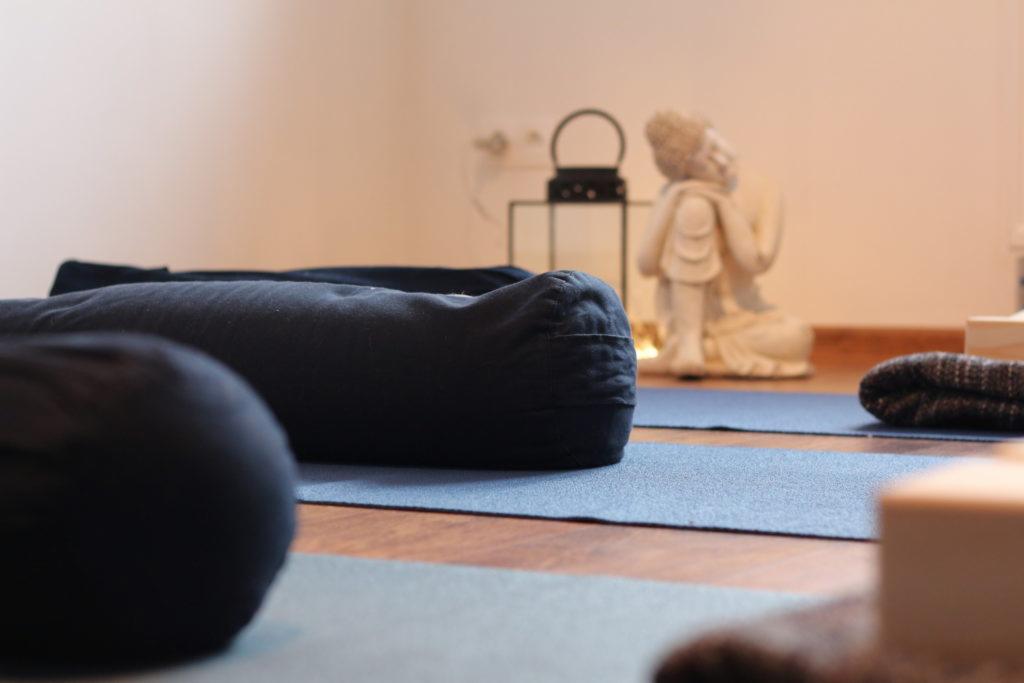 Studio jogi: Poduszki do medytacji na niebieskich matach, drewniana podłoga, statuetka Buddy i lampion w tle.