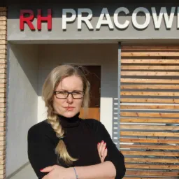 Portret kobiety z blond warkoczem na tle budynku z napisem 'RH PRACOWNIA', ceglaną ścianą i drewnianą przesłoną.
