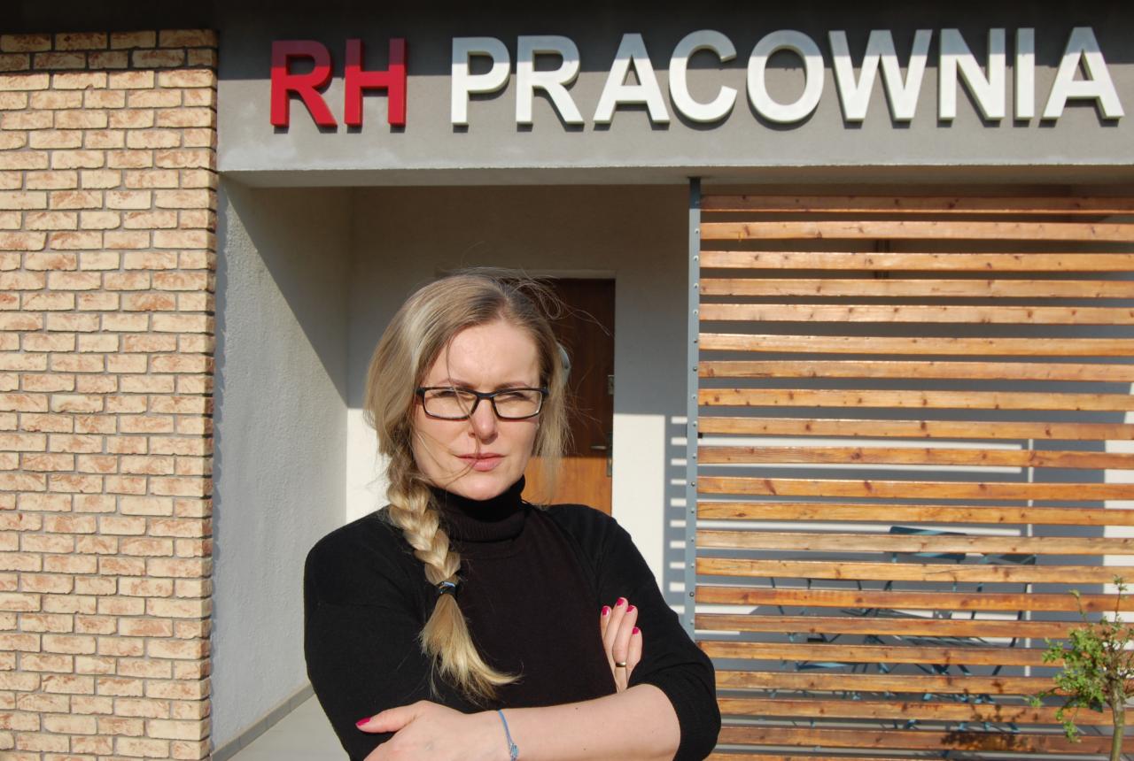 Portret kobiety z blond warkoczem na tle budynku z napisem 'RH PRACOWNIA', ceglaną ścianą i drewnianą przesłoną.