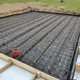 Przygotowanie zbrojenia płyty fundamentowej: siatka stalowa z dystansami na folii, w szalunku z drewnianych belek, widoczny pomarańczowy odpowietrznik w rogu, w tle domy jednorodzinne i trawa.