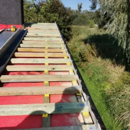 Konstrukcja dachu z łatami i kontrłatami zamocowanymi żółtymi elementami dystansowymi, widoczne elementy komina, narzędzia i krajobraz w tle.