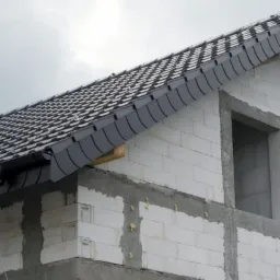 Fragment budowanego domu z białego bloczka z zamontowanym, grafitowym dachem z dachówki karpiówki i orynnowaniem, widoczne drewniane elementy konstrukcyjne pod okapem.