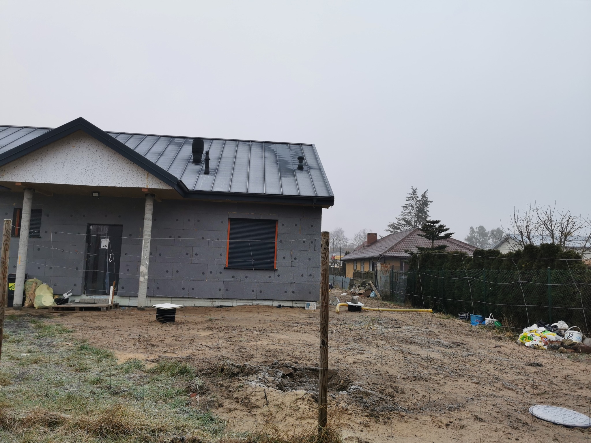 Surowy dom szkieletowy w trakcie budowy, ocieplony styropianem, z zamontowanym, błyszczącym, metalowym dachem na tle pochmurnego nieba.