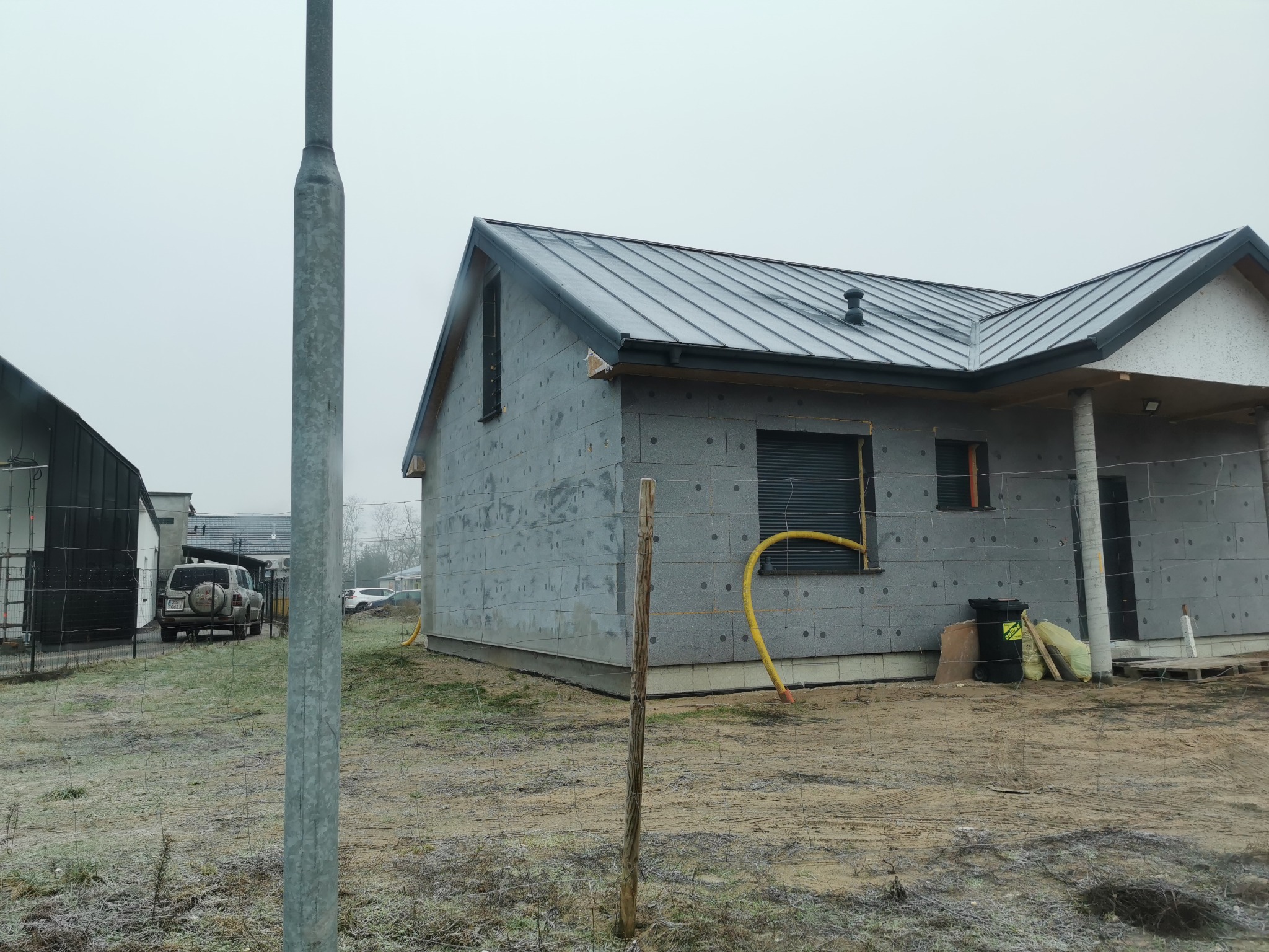 Surowy stan domu jednorodzinnego z szarej bloczkowej elewacji, z ciemnym dachem, widoczny żółty wąż doprowadzający media, teren budowy bez trawy, ogrodzony siatką.