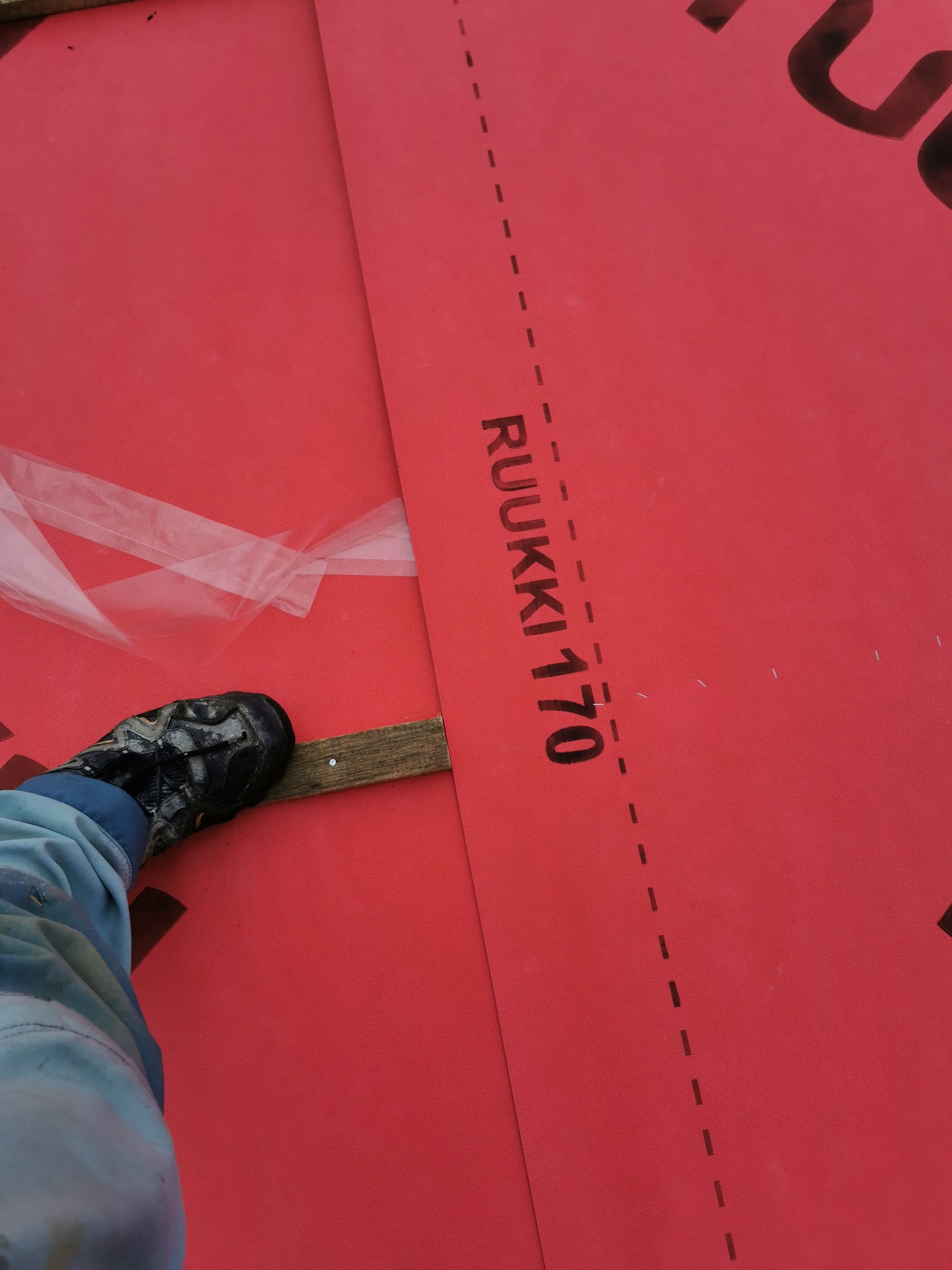 Fragment czerwonej membrany dachowej Ruukki 170 z widoczną stopą w bucie roboczym i fragmentem deski podczas montażu.