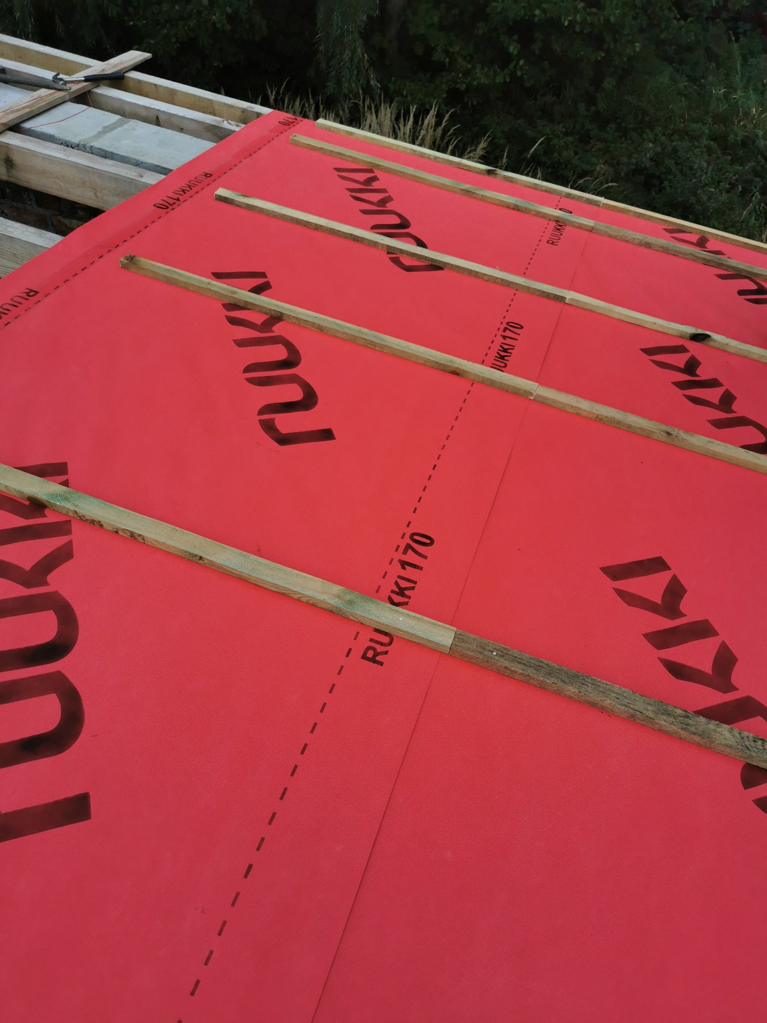 Czerwona membrana dachowa z czarnym nadrukiem producenta, zabezpieczona drewnianymi łatami, widok z góry podczas montażu.