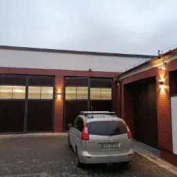Budynek z bramami garażowymi z cegły klinkierowej oświetlony zewnętrznymi lampami ściennymi, widoczna instalacja odgromowa na dachu, zaparkowany samochód osobowy.