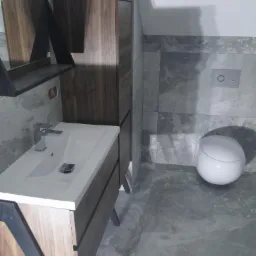 Nowoczesna łazienka z szarymi płytkami imitującymi kamień, wisząca toaleta i umywalka z szafką w odcieniach brązu i czerni.