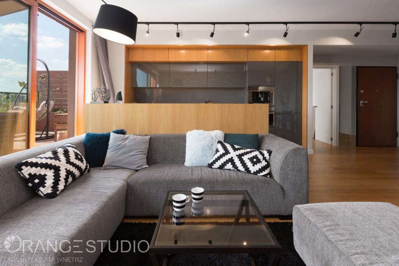 Szary narożnik z wzorzystymi poduszkami i stolikiem kawowym na tle otwartej kuchni z drewnianymi i szarymi frontami, widok na taras z huśtawką.