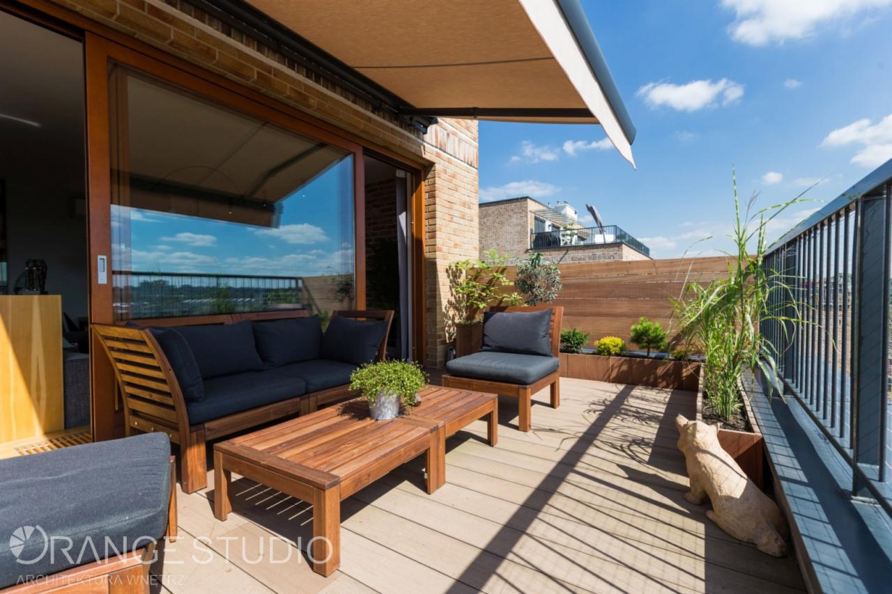 Drewniany taras z sofą, fotelem i stolikiem kawowym, osłonięty markizą, z widokiem na miasto i niebo z chmurami; na podłodze dekoracyjna rzeźba kota.