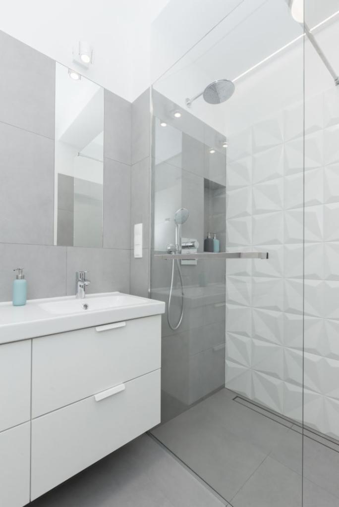 Nowoczesna łazienka w odcieniach szarości i bieli, z umywalką nablatową na szafce z szufladami, przeszklonym prysznicem typu walk-in i dekoracyjną ścianą z trójwymiarowych płytek.