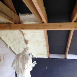 Prace izolacyjne poddasza: aplikacja jasnej piany izolacyjnej między krokwiami dachu, widoczna osoba w kombinezonie ochronnym podczas natrysku.